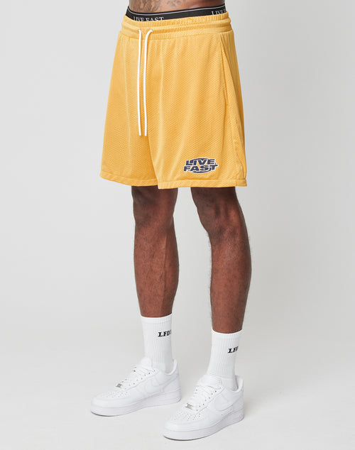 A person wears yellow LFDY Oval Mesh Shorts by LIVE FAST DIE YOUNG, featuring an elastic waistband and LIVE FAST text, paired with white socks with black LFDY text and white sneakers, standing against a plain white background.