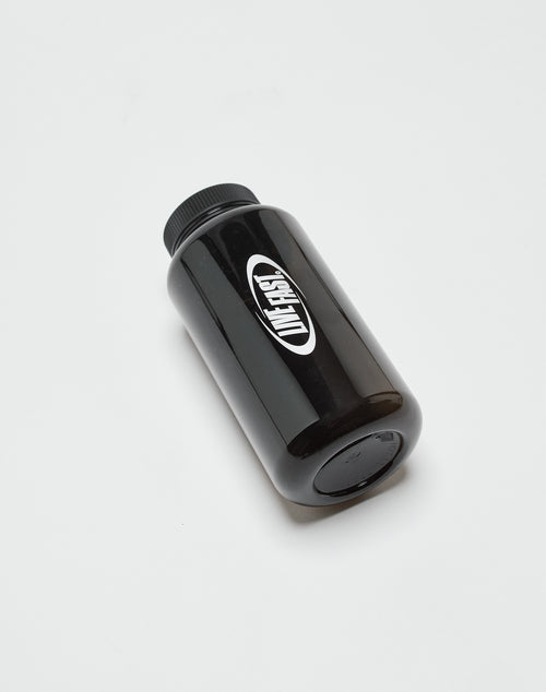 A black plastic Live Fast Performance Gym Bottle from LIVE FAST DIE YOUNG, featuring a black screw cap, lies on its side on a white surface. The BPA-free bottle has a white oval label with bold black branding.