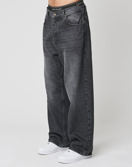 A person wearing LFDY Baggy Denim by LIVE FAST DIE YOUNG—baggy, faded black jeans with a wide straight leg made from 100% cotton—and white sneakers stands against a plain white background. The waistband reads LIVE FAST.