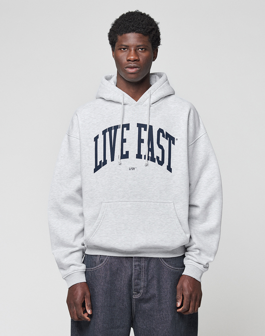A young man faces the camera in a light gray Live Fast College Hooded sweatshirt by LIVE FAST DIE YOUNG, featuring a bold "LIVE FAST" front print, paired with dark jeans against a plain light gray background.