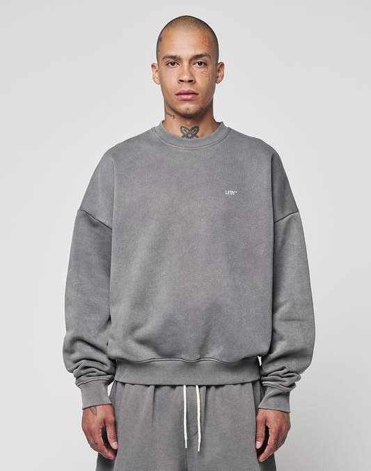 A person with a shaved head and tattoos wears the LIVE FAST DIE YOUNG Basic Chest Sweater—an oversize fit, faded gray sweatshirt made from soft Baumwolle—along with matching sweatpants, against a plain light gray background.