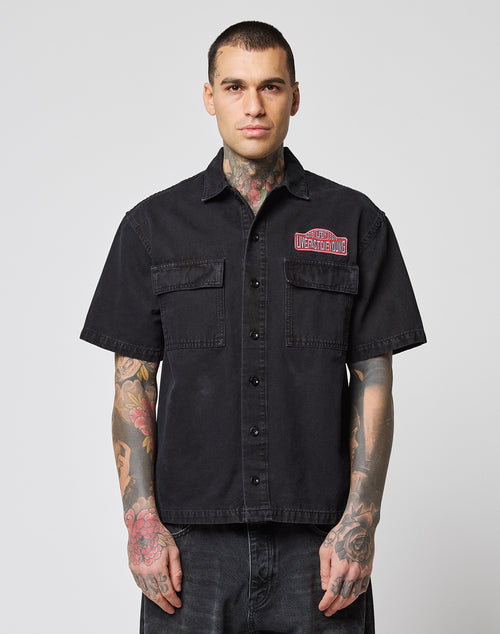 A man with tattoos models the LFDY Workwear Shirt by LIVE FAST DIE YOUNG, a black oversized cotton short-sleeve button-up with two chest pockets and a red patch on the left, standing facing forward against a plain light background.