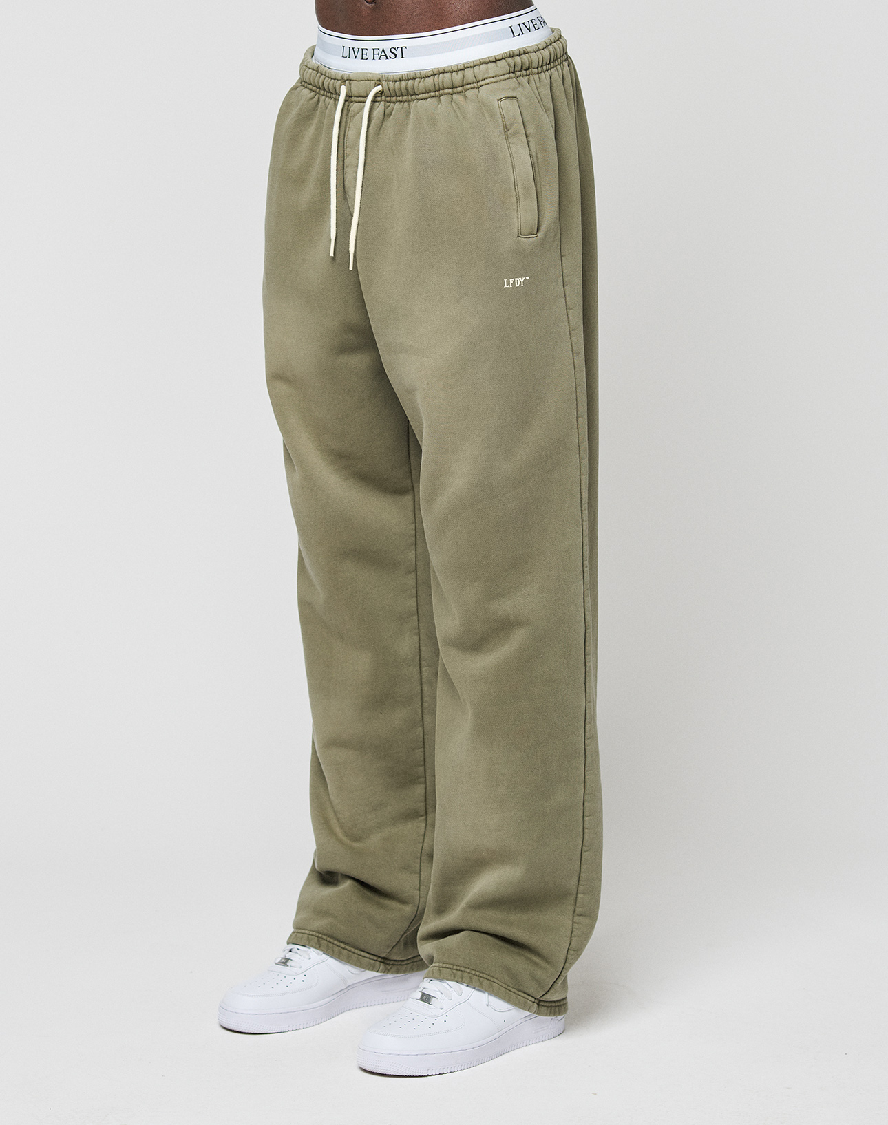 A person wears the LIVE FAST DIE YOUNG Basic Wide Jogger—olive green, wide fit, with “LIVE FAST” on the waistband and left pocket—paired with white sneakers, standing against a plain white background.