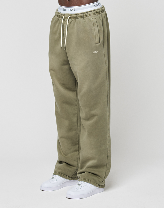 A person wears the LIVE FAST DIE YOUNG Basic Wide Jogger—olive green, wide fit, with “LIVE FAST” on the waistband and left pocket—paired with white sneakers, standing against a plain white background.