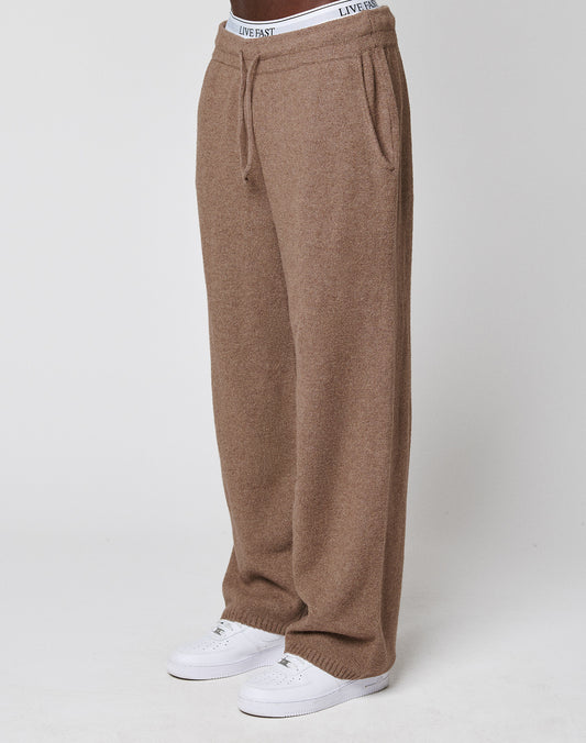 A person wears the LIVE FAST DIE YOUNG Live Fast Boucle Pants in loose-fitting brown knit with an elastic waistband and straight leg, paired with white sneakers and a visible "LIVE FAST" waistband, against a plain white background.