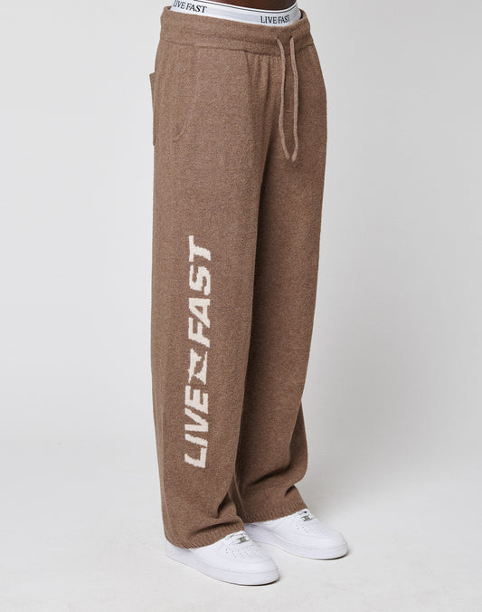 A person wears LIVE FAST DIE YOUNG's Live Fast Boucle Pants, wide fit and brown with white "LIVE FAST" on the left leg and elastic waistband. Styled with white sneakers against a plain light background.