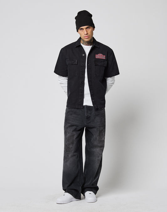 A person wears a black LFDY Workwear Shirt by LIVE FAST DIE YOUNG over a white long-sleeve, loose vintage black jeans, black beanie, and white sneakers, standing against a plain white background.