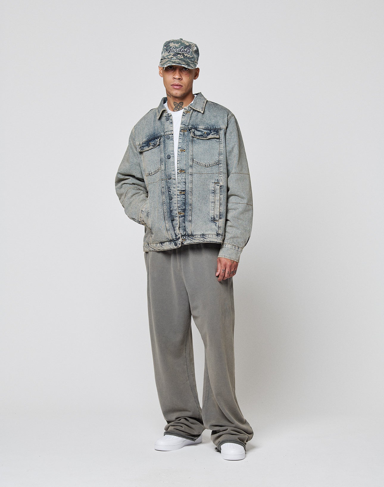 A man stands against a plain background wearing the LIVE FAST DIE YOUNG Puffed Denim Coach Jacket in light blue, gray baggy pants, white shirt, white sneakers, and a patterned cap, with his hands tucked into his jacket pockets.