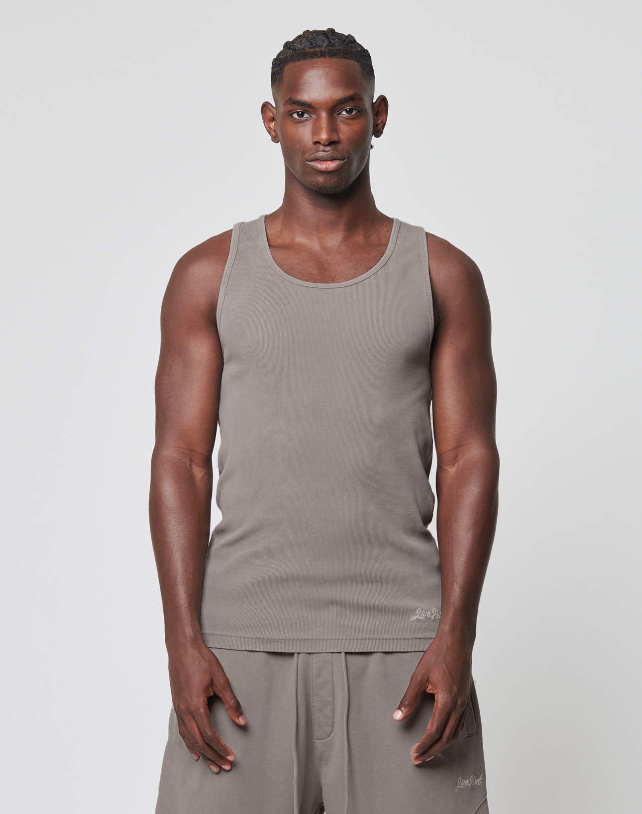 A man with short hair stands facing forward against a plain white background, wearing the LIVE FAST DIE YOUNG Basic Rib Tank in light brown and matching shorts, his arms relaxed at his sides and a neutral expression on his face. | model