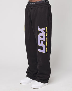 A person wears the LFDY Racing Wide Jogger by LIVE FAST DIE YOUNG, featuring bold white and yellow leg prints, a wide fit, and a broad elastic waistband, paired with white sneakers against a plain light background.