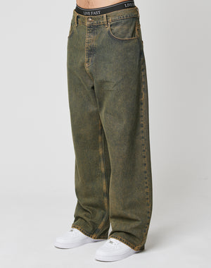 Someone wears LIVE FAST DIE YOUNG’s LFDY Baggy Denim—baggy, faded greenish-brown jeans with wide straight legs and front pockets, made of 100% cotton—showing their black LIVE FAST waistband above the jeans, paired with white sneakers.