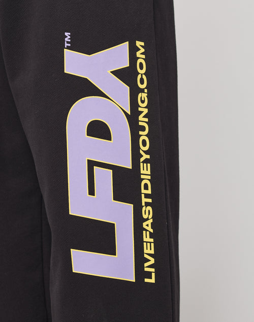 A close-up of black Baumwolle fabric shows large purple LFDY letters outlined in yellow with LIVEFASTDIEYOUNG.COM printed vertically. The LFDY Racing Wide Jogger by LIVE FAST DIE YOUNG features a wide fit and elastic waistband for comfort and style.