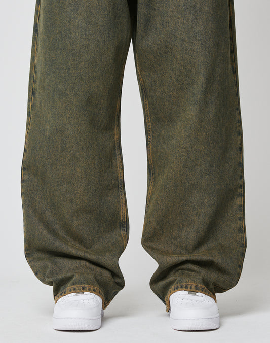 A person wears LFDY Baggy Denim by LIVE FAST DIE YOUNG—baggy fit, faded green jeans with a wide straight leg made from 100% cotton—bunching at the ankles over white sneakers on a plain white background.