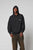 A person wears a black LFDY Hardwear Hooded by LIVE FAST DIE YOUNG with the hood up and chest logo, paired with loose-fitting faded brown Oversize Fit pants, standing against a plain light gray background. The hoodie is 100% Baumwolle.