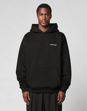 A person in the Live Fast Plain Hooded by LIVE FAST DIE YOUNG—a black, oversized cotton-polyester hoodie with LIVE FAST in white on the left chest—stands against a plain light gray background.