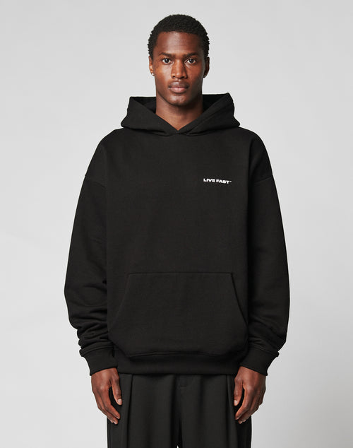 A person in the Live Fast Plain Hooded by LIVE FAST DIE YOUNG—a black, oversized cotton-polyester hoodie with LIVE FAST in white on the left chest—stands against a plain light gray background.