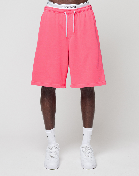 A person in bright pink Basic Shorts by LIVE FAST DIE YOUNG, with an elastic waistband, white socks, and white sneakers stands against a plain white background. The waistband displays the LIVE FAST branding on their underwear.