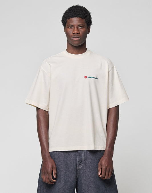 A person stands against a plain background wearing the LIVE FAST DIE YOUNG LFDY Dealership Tee—an oversize, off-white 100% Baumwolle T-shirt with a small red and blue LIVEFAST logo on the left chest—paired with dark wide-leg jeans.