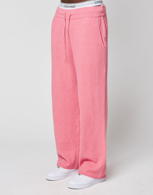 A person wears loose-fitting, wide fit pink Live Fast Boucle Pants by LIVE FAST DIE YOUNG, featuring an elastic waistband with "LIVE FAST" text. Styled with white sneakers against a plain, light background.