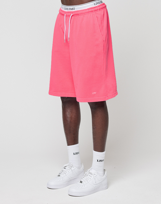 A person wears LIVE FAST DIE YOUNG Basic Shorts in bright pink with an elastic waistband, white socks with “LEDY” text, and white Nike sneakers. Only the lower half of their body is visible against a plain white background.