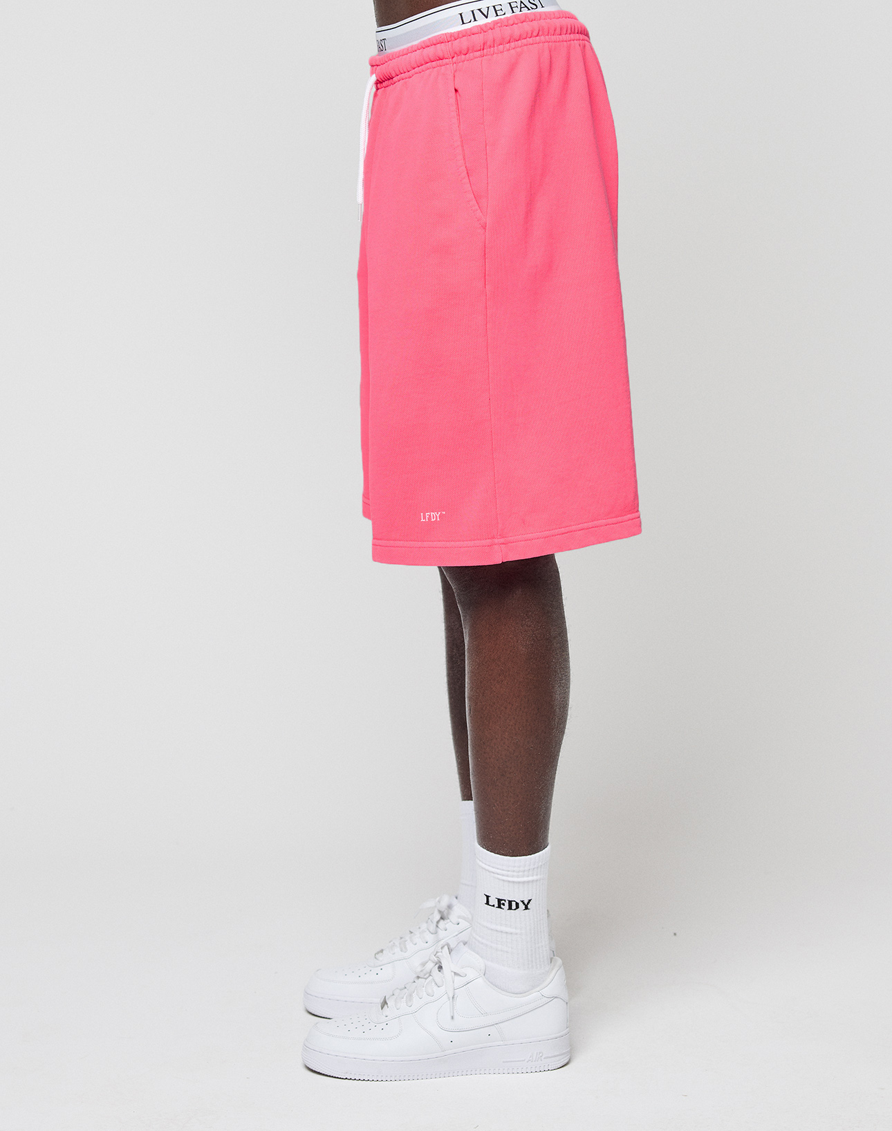 A person stands sideways in bright pink Basic Shorts by LIVE FAST DIE YOUNG, white logo-print waistband underwear, white socks, and white Nike sneakers against a plain white background.