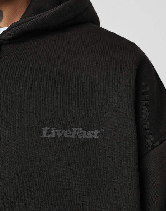 A close-up of a person wearing the LIVE FAST DIE YOUNG Basic 510 Hooded Zip in black with Vintage-Waschung and a subtle gray LiveFast chest print. The oversize fit highlights the neck tattoo of the wearer.