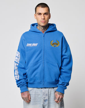 A man wears the LFDY Team Hooded Zip by LIVE FAST DIE YOUNG, featuring an oversized fit, blue color, yellow crest, LIVE FAST text, white sleeve detail, and Flaggen-Stick accents. He stands forward with tattoos on his hands and neck visible.