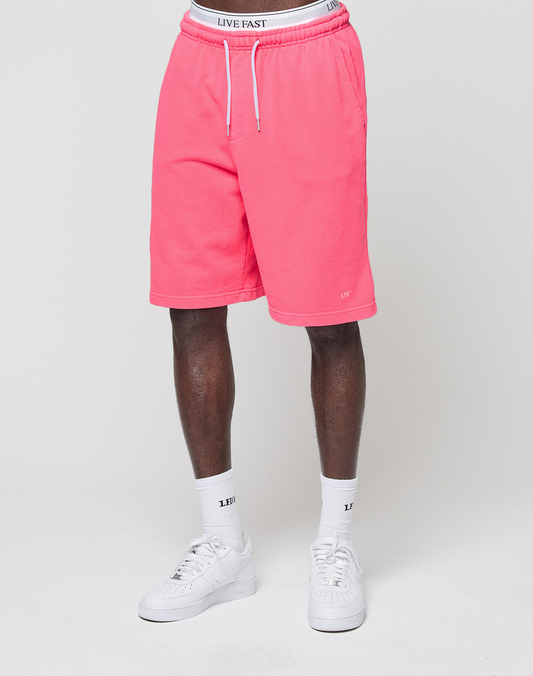 A person in bright pink LIVE FAST DIE YOUNG Basic Shorts with elastic waistband, white "LIVE EAST" socks, and white sneakers stands against a plain light background. The upper body is not visible.