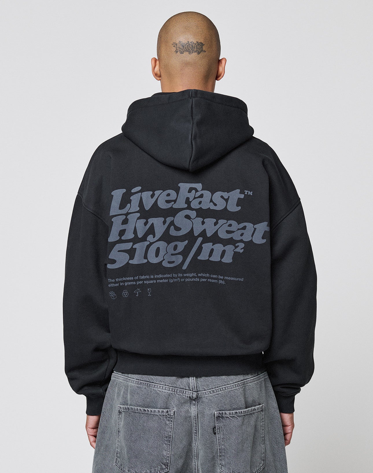 A person with a shaved head and a tattoo on the back of their scalp wears the LIVE FAST DIE YOUNG Basic 510 Hooded Zip in Oversize Fit with a large gray Backprint text, paired with gray jeans.
