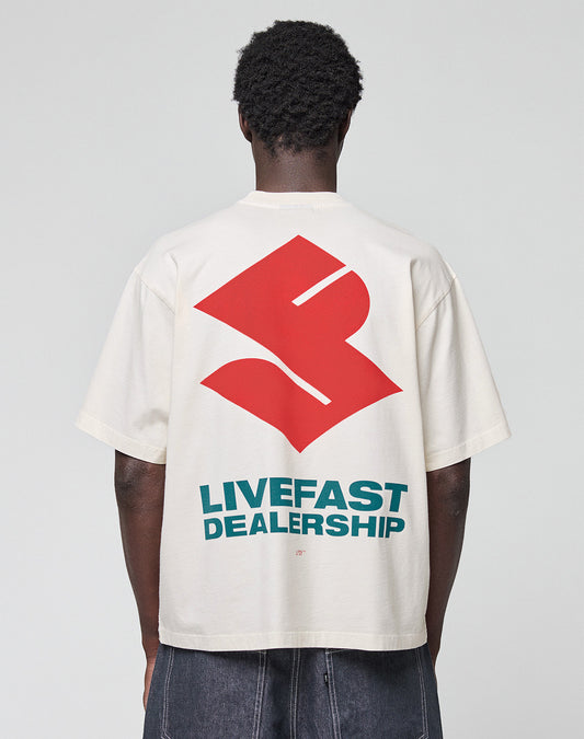 A person faces away, wearing the LFDY Dealership Tee by LIVE FAST DIE YOUNG—an oversize white 100% Baumwolle shirt with a bold red abstract backprint and "LIVEFAST DEALERSHIP" in striking teal letters.