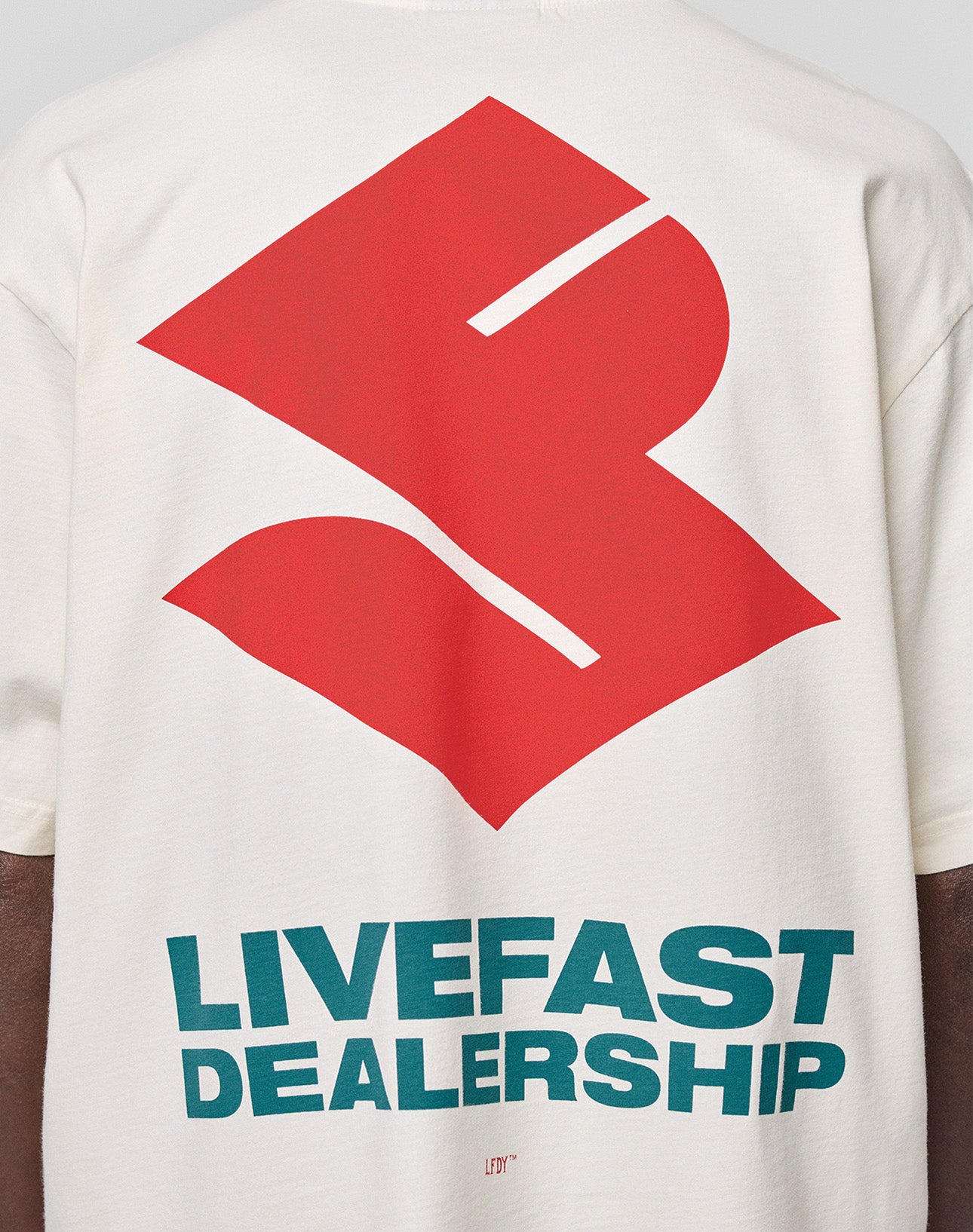 A person wears the LFDY Dealership Tee by LIVE FAST DIE YOUNG, an oversized white 100% Baumwolle shirt with a bold red abstract back graphic and teal "LIVEFAST DEALERSHIP" text, featuring striking front and back prints.