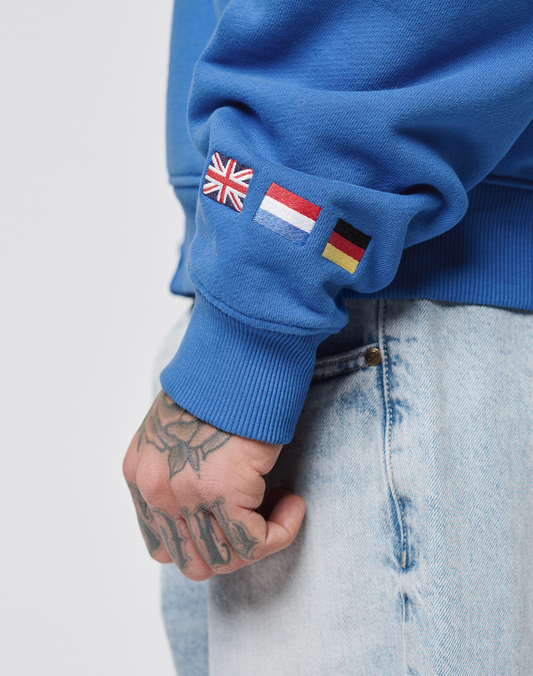 A person wears light denim jeans and the LFDY Team Hooded Zip by LIVE FAST DIE YOUNG in Oversize Fit, featuring multilogo embroidery plus UK, Netherlands, and Germany flag patches on the sleeve. Unique tattoos are visible on their hand.