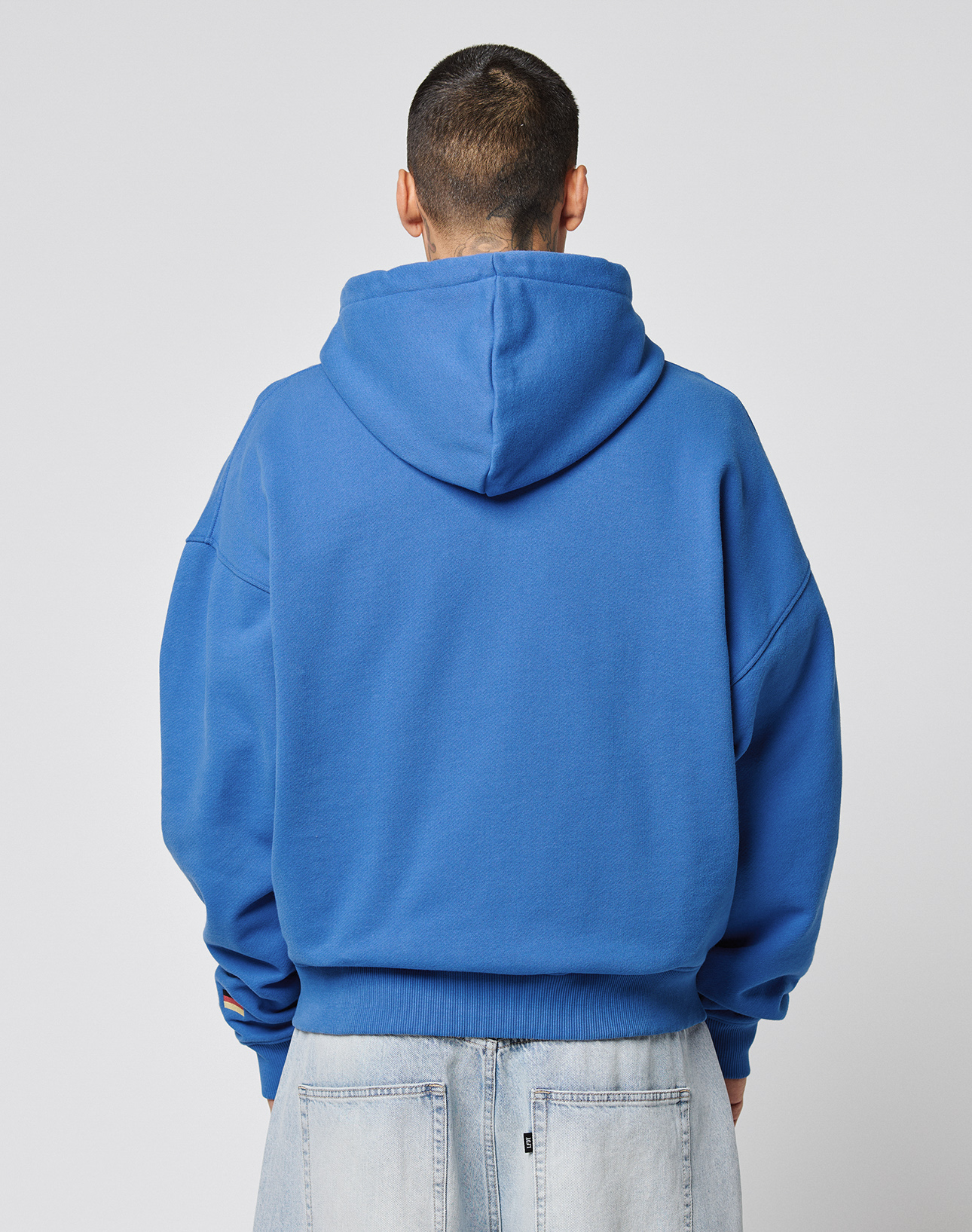 A person stands facing away from the camera, wearing an oversize fit bright blue LFDY Team Hooded Zip by LIVE FAST DIE YOUNG with Multilogo Stickereien and light blue jeans against a plain light background.