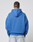 A person stands facing away from the camera, wearing an oversize fit bright blue LFDY Team Hooded Zip by LIVE FAST DIE YOUNG with Multilogo Stickereien and light blue jeans against a plain light background.