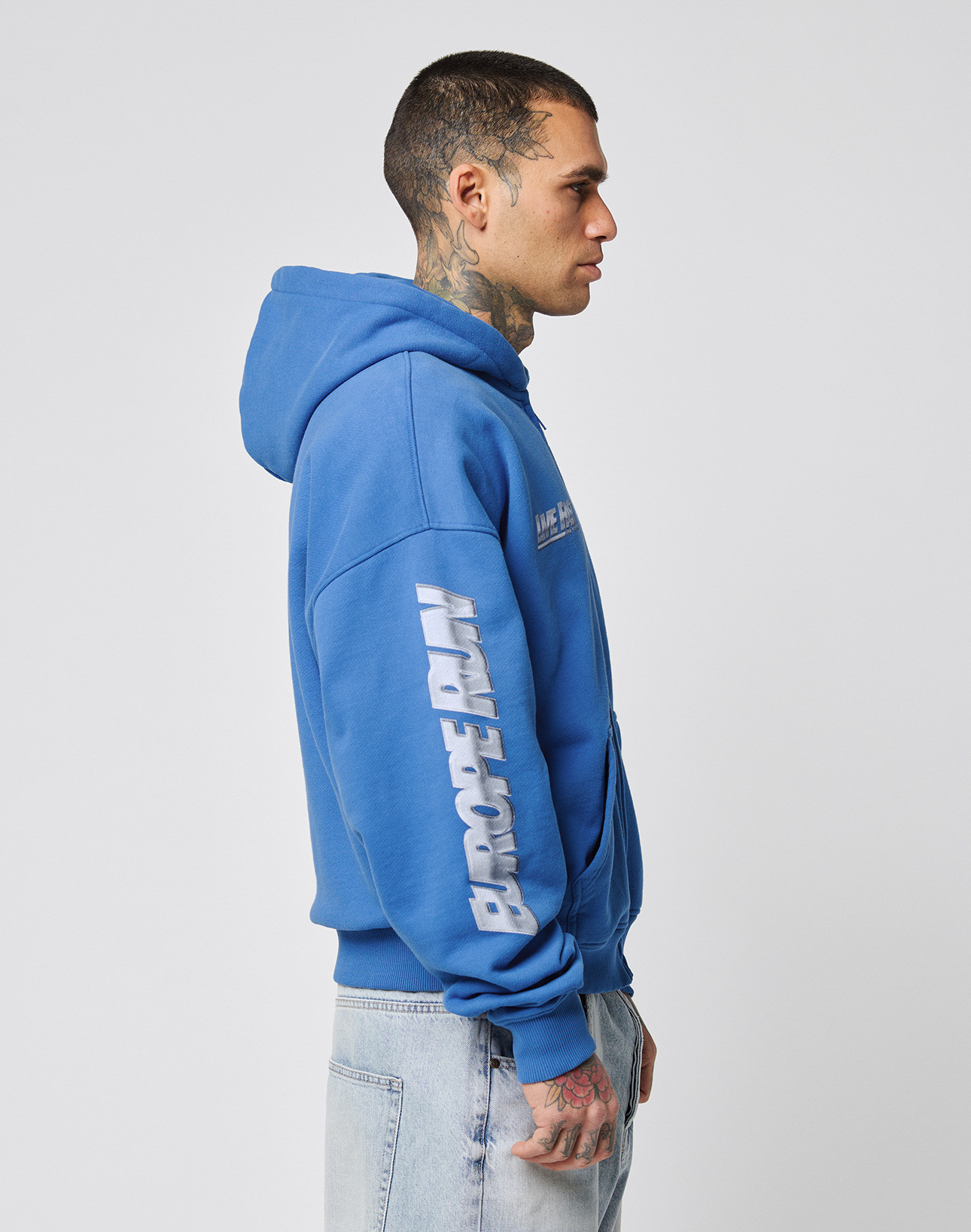 A tattooed person in profile wears the LIVE FAST DIE YOUNG LFDY Team Hooded Zip—an oversize blue hoodie with EUROPE RUN and multilogo Stickereien on the sleeve—paired with light blue jeans, set against a plain white background.