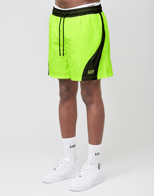 A person wears LIVE FAST DIE YOUNG Live Fast Nylon Shorts in neon yellow with black accents, logo on the left leg, elastic waist with black drawstring, paired with white socks and sneakers, standing against a plain white background. | model