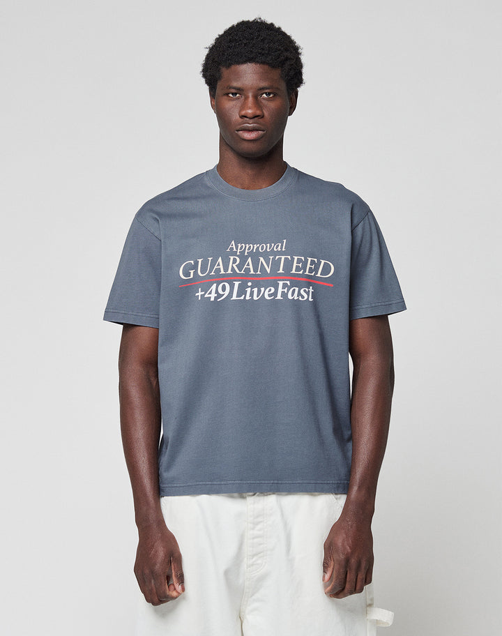A young man wears the LFDY Guaranteed Tee by LIVE FAST DIE YOUNG, a regular fit gray t-shirt made of 100% cotton with bold “Approval GUARANTEED +49LiveFast” front print, paired with white pants against a light gray background.