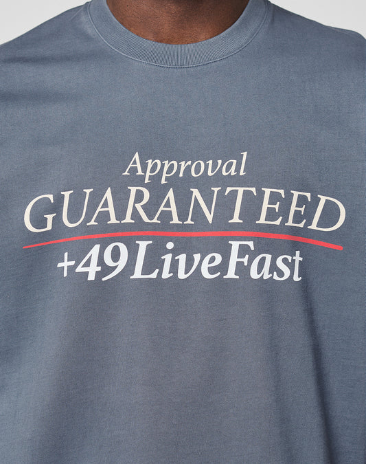 A person wearing the LFDY Guaranteed Tee by LIVE FAST DIE YOUNG—a regular fit blue shirt in 100% cotton, with a bold white and red “Approval GUARANTEED +49LiveFast” front print.