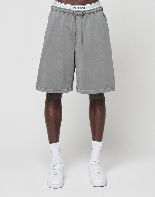 A person wears LIVE FAST DIE YOUNG Basic Shorts in regular fit gray with a faded finish and a white waistband reading "LIVE FAST," paired with white socks and sneakers, standing against a plain white background. Upper body not shown.