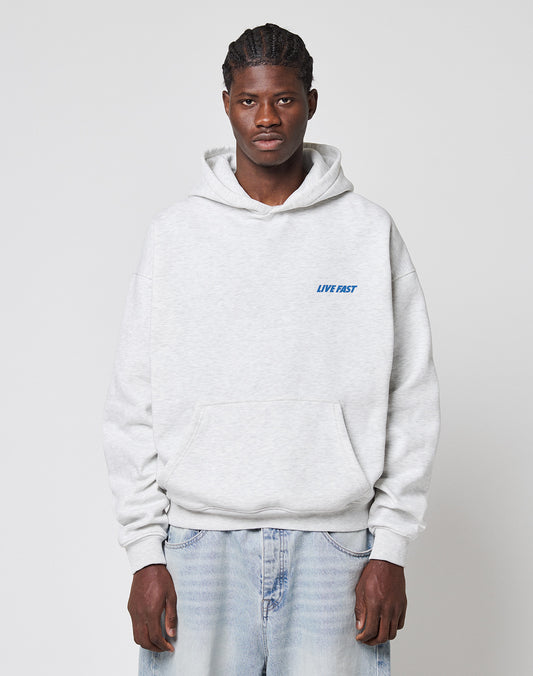 A person with braided hair wears the LFDY Globe Hooded by LIVE FAST DIE YOUNG, an oversize light gray Baumwolle Polyester hoodie with LIVE FAST in blue on the left chest, and light blue jeans, standing against a plain white background.