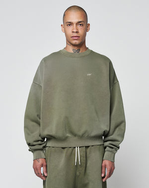 A person wears the LIVE FAST DIE YOUNG Basic Chest Sweater, an oversize fit olive green sweatshirt with subtle lettering and a handgefertigter Fade-Effekt, paired with matching drawstring sweatpants against a light gray background.