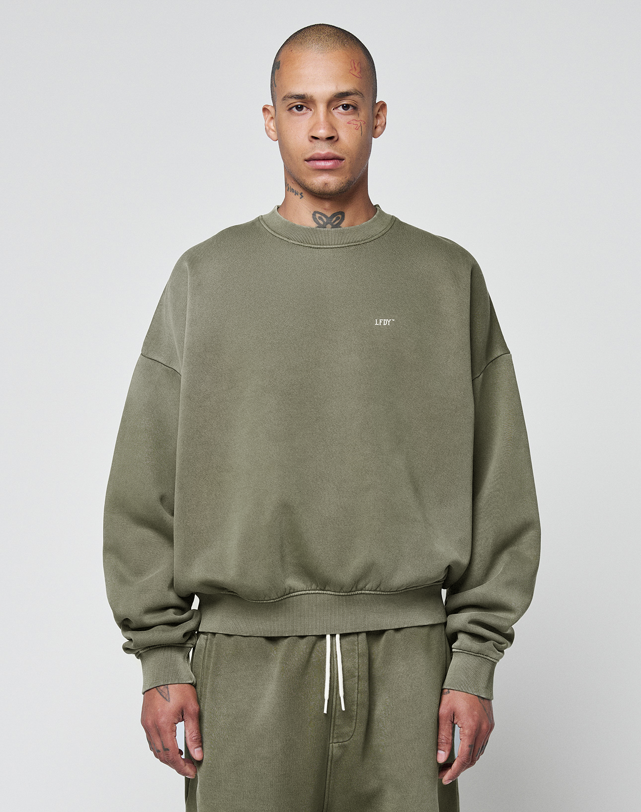 A person wears the LIVE FAST DIE YOUNG Basic Chest Sweater in olive green with a handmade fade effect and matching sweatpants, standing facing forward against a light gray background. The oversize fit sweater features small chest text.