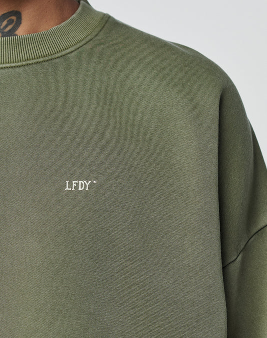 Close-up of a person wearing the LIVE FAST DIE YOUNG Basic Chest Sweater in olive green, featuring a subtle hand-finished fade effect, small white LFDY⢠chest text, and an Oversize Fit that reveals part of the neck and a visible tattoo.