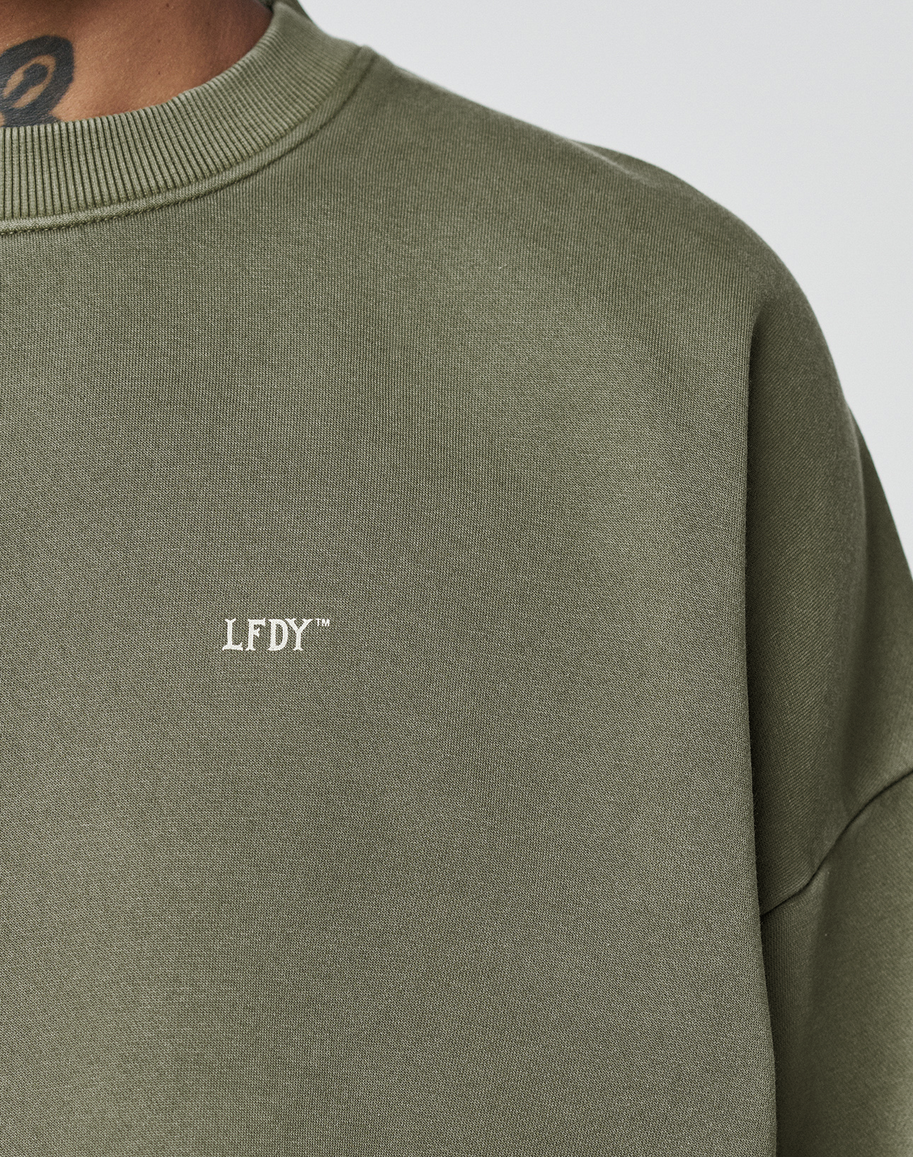 Close-up of a person wearing the LIVE FAST DIE YOUNG Basic Chest Sweater in olive green with white LFDY™ text on the chest and an Oversize Fit. The neck, part of the shoulder, and a neck tattoo are visible.