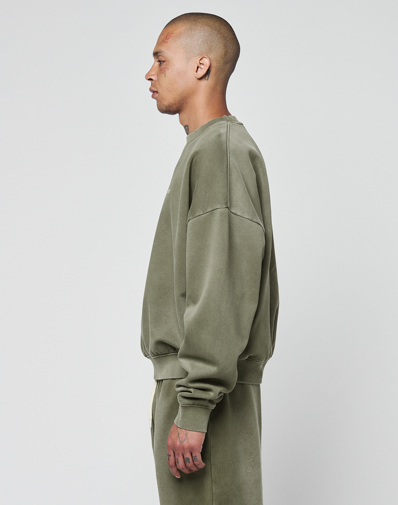 A person with a shaved head stands in profile, wearing an olive green LIVE FAST DIE YOUNG Basic Chest Sweater and matching sweatpants in oversize fit, against a plain white background.