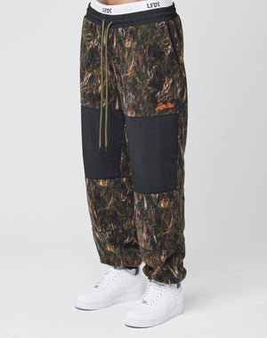A person wears LFDY Fleece Pants by LIVE FAST DIE YOUNG, featuring camo print, black knee patches, nylon panels, Leisure Paris orange embroidery, drawstring waist, elastic cuffs, and an oversized fit—paired with white sneakers.