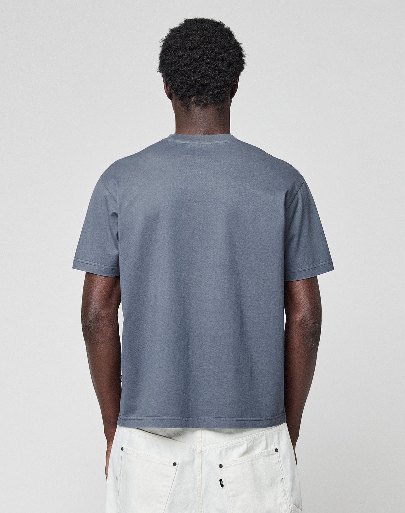 A person faces away from the camera, wearing the LIVE FAST DIE YOUNG LFDY Guaranteed Tee, a plain gray short-sleeve t-shirt in 100% Baumwolle, paired with light-colored pants against a plain light background.