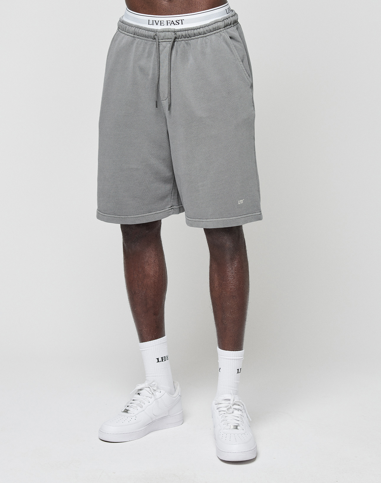 A person models LIVE FAST DIE YOUNG Basic Shorts in grey with an elastic waistband, paired with LFT logo white socks and white sneakers, standing against a plain white background. Only their lower torso and legs are visible.