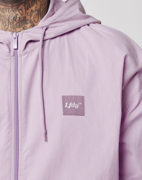 A person wears the Basic Light Hooded Zip by LIVE FAST DIE YOUNG, a light purple, regular fit jacket with zipper and oversized hood. A square chest patch reads Lfdv™; text tattoos are visible on the persons neck.