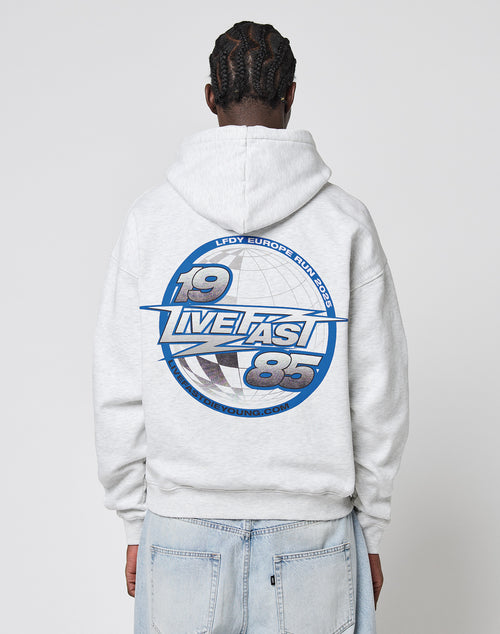 A person with braided hair is wearing the LFDY Globe Hooded by LIVE FAST DIE YOUNG—an oversize light gray hoodie. The back has a bold blue and white 19 LVETASY 85 and LFDY Europe Run 2023 graphic. The person faces away from the camera.
